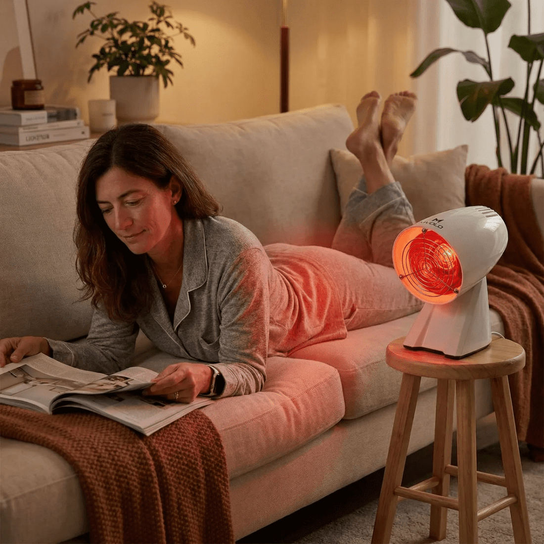 Vrouw ligt ontspannen op de bank en leest een tijdschrift terwijl een infrarood warmtelamp haar voeten en benen verwarmt in een gezellige woonkamer