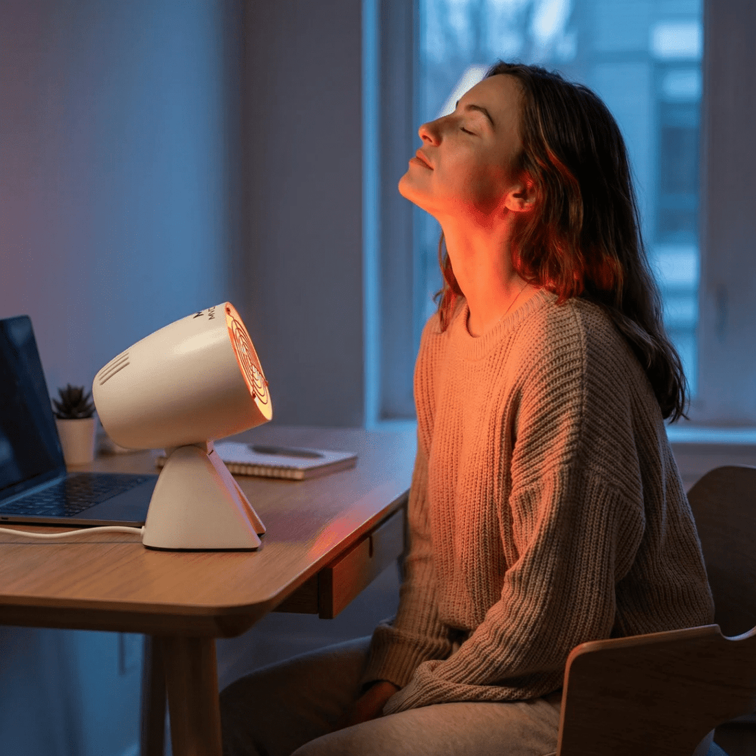 Vrouw gebruikt een infraroodlamp voor het gezicht aan bureau, warm licht voor ontspanning en huidroutine